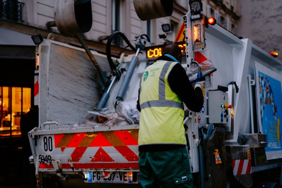 A waste collection worker wearing a high-visibility yellow vest with grey reflective stripes and green trousers is operating a garbage truck on a city street during dusk or early evening. The worker is facing away from the camera, with a blue glove on their right hand as they tend to the truck's controls or paperwork. The garbage truck has a white exterior with red and orange reflective safety markings on its rear, including diagonal stripes and warning lights. The open rear compartment of the truck is filled with black and grey mixed waste, slightly spilling over the edge, and appears to be in the process of processing rubbish for disposal. In the background, a building with illuminated windows and exterior wall textures can be seen, indicating an urban environment. The scene captures a typical private waste collection activity, highlighting independent rubbish removal operations often used for bulky or non-standard waste, consistent with services like those provided by Rubbish Clearance Mayfair in the W1J postcode area.