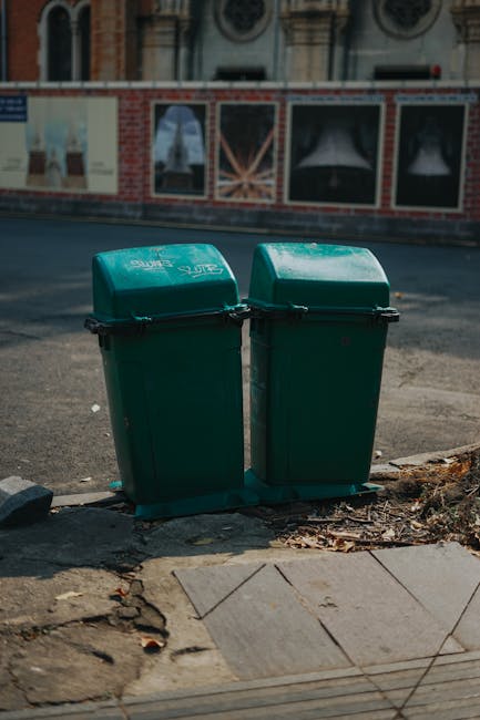 Two green plastic wheelie bins with slightly domed lids and black handles are positioned side by side on a paved sidewalk near the edge of an asphalt road. The bins show minor surface scuffs and are marked with white branding or text, indicating they are for waste collection. Behind the bins, a brick and brick-patterned wall features colorful posters or advertisements with images and text, some of which include window-like sections. The setting appears to be an urban street environment with a building façade in the background, partially visible above the wall, showcasing arched windows and decorative architectural details. The scene is illuminated by natural daylight, creating soft shadows on the ground and highlighting the textures of the paving, the wall, and the containers. The placement of the bins suggests they are set out for collection, consistent with private waste disposal and rubbish removal services like those offered by Rubbish Clearance Mayfair, serving the local Mayfair W1J area. The environment overall aligns with typical urban waste management practices involving independent rubbish collection points on residential or commercial streets.
