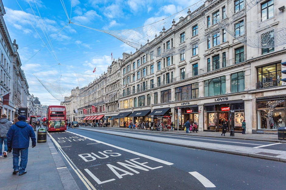 A busy street scene in Mayfair with a row of multi-storey white stone buildings housing retail stores, including a Levi's shop with large glass windows displaying sale signs. Pedestrians walk along the wide pavement, some pushing strollers or carrying shopping bags. A red double-decker bus is stopped at a bus stop marked 'BUS STOP' on the dark asphalt road, which features designated bus lane markings. Overhead, a network of wires is strung between buildings, likely for trolley or tram services. The sky above is bright with scattered clouds, illuminating the scene with natural daylight. The storefronts exhibit modern signage with contrasting black and red backgrounds. The image depicts an urban retail environment typical of central London, highlighting commercial activity and pedestrian traffic, with the potential context of private waste or rubbish clearance services servicing retail premises like those visible in the photograph.