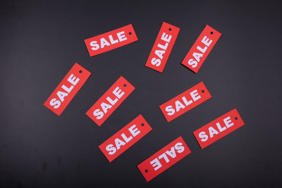 Multiple red rectangular sale tags with white bold lettering spelling out 'SALE' are scattered across a dark, smooth surface. The tags have small black holes at the top left corners, indicating they are designed for hanging or attachment. The surface appears to be matte with no visible texture, providing a stark contrast to the bright red tags. The arrangement of the tags seems random, with some overlapping slightly and others spaced apart, creating a dynamic visual composition. The lighting is even, without harsh shadows, emphasizing the clarity of the printed text. Although the scene does not involve rubbish or removal activities directly, the image could be relevant in a retail context, such as shop clearance or private disposal preparations where sale signs might be used for clearance items, aligning with independent waste handling or clearance services like those offered by Rubbish Clearance Mayfair in the context of retail and commercial waste disposal scenarios.