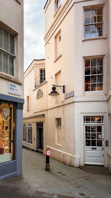 A busy street scene in Mayfair with a row of multi-storey white stone buildings housing retail stores, including a Levi's shop with large glass windows displaying sale signs. Pedestrians walk along the wide pavement, some pushing strollers or carrying shopping bags. A red double-decker bus is stopped at a bus stop marked 'BUS STOP' on the dark asphalt road, which features designated bus lane markings. Overhead, a network of wires is strung between buildings, likely for trolley or tram services. The sky above is bright with scattered clouds, illuminating the scene with natural daylight. The storefronts exhibit modern signage with contrasting black and red backgrounds. The image depicts an urban retail environment typical of central London, highlighting commercial activity and pedestrian traffic, with the potential context of private waste or rubbish clearance services servicing retail premises like those visible in the photograph.