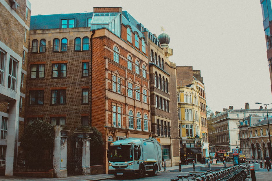 The image depicts a street scene in an urban area with multiple historic brick and stone buildings featuring large, arched windows and ornate architectural details. The primary focus is a white refuse collection truck parked on the right side of the street, positioned in front of a narrow sidewalk with black bollards. The truck appears to be designed for waste removal or rubbish collection, with a compact cab and a large rear compartment for waste storage. Behind the truck, there is a small gated entrance with brick and stone pillars leading to a private property, possibly a courtyard or residence. The surrounding environment includes other multicoloured buildings, with facades painted in pastel shades and intricate window treatments, consistent with a city centre setting. The street is lined with additional parked vehicles and a few bicycles secured to a bike rack. Overhead, street lamps and signage are visible, contributing to the typical urban streetscape. Natural daylight reveals a clear sky, emphasizing the clean and organized appearance of this private waste collection scene, subtly aligned with independent rubbish removal services such as those offered by Rubbish Clearance Mayfair for alternative waste handling of bulky items and urban rubbish. 