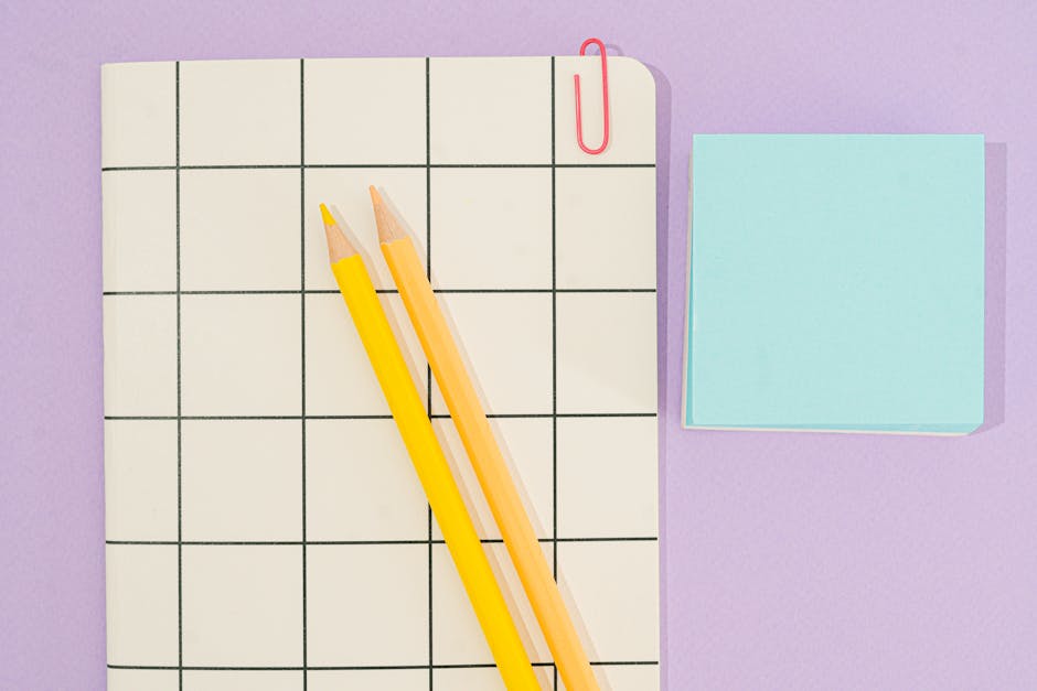 A flat-lay image features a white, grid-patterned notepad placed on a light purple surface, with two yellow pencils lying diagonally across the notepad. To the right of the notepad, there is a small stack of light blue sticky notes. A red paperclip clips the top right corner of the notepad. The scene is well-lit with soft, even lighting, creating a clean and organized workspace impression. The neutral tones and simple objects suggest planning or note-taking related to waste management or rubbish removal tasks, aligning subtly with the context of private or independent rubbish collection services provided by Rubbish Clearance Mayfair.