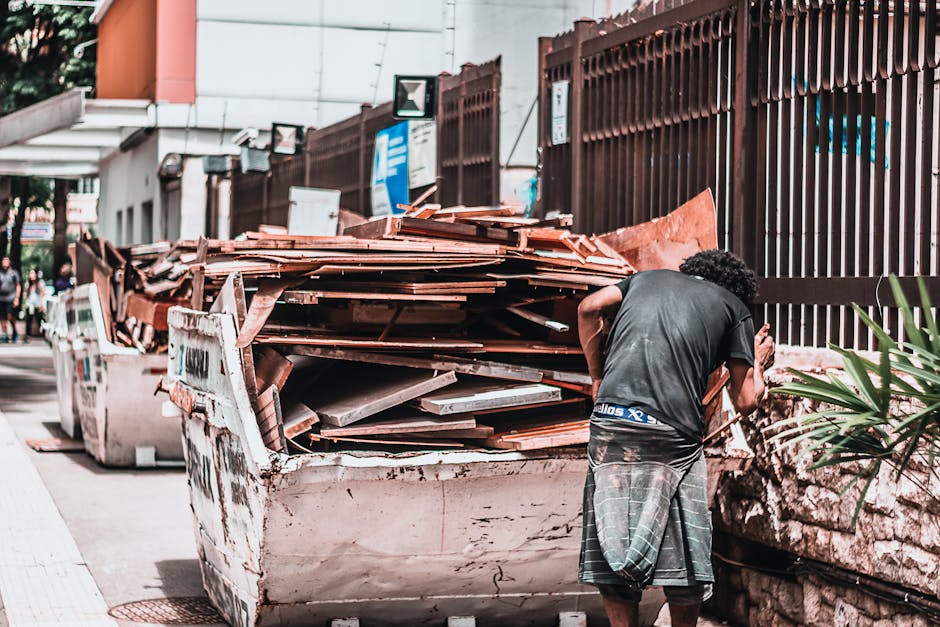 A man with curly dark hair, dressed in a black t-shirt and plaid shorts, is seen standing next to a large, white, industrial skip bin filled with a variety of construction debris, including wooden planks, tiles, and broken materials. The wooden planks are reddish-brown with a rough, splintered texture, while the tiles appear grey and broken, with some pieces stacked unevenly. The skip bin, which shows signs of wear and rust, is positioned on a paved sidewalk next to a tall, brown metal fence with vertical slats. In the background, there are blurred images of a building with modern architecture, street signs, and a few pedestrians. The scene is captured in natural daylight, providing clear visibility of the materials and environmental context, illustrating typical on-site waste removal associated with private disposal or alternative rubbish collection methods.