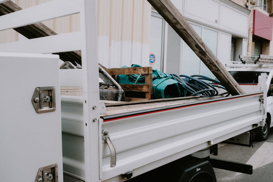 A white flatbed utility truck parked on a city street with an open cargo area loaded with various items for rubbish removal, including a wooden pallet, a coil of black industrial hose, and a green strap or cover. The truck is positioned near a building with large glass windows and metal panels, indicating an urban environment. The cargo area extends to the right side of the image, with the tailgate open, showing the contents arranged for transport. The scene appears to be part of a private waste disposal or on-site clearance operation, typical of independent collection services. The overall atmosphere is neutral and professional, emphasizing the logistical aspect of rubbish collection in a residential or commercial setting.