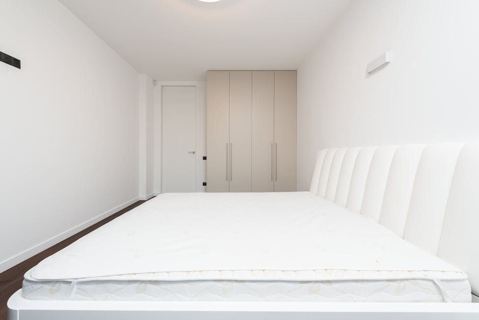 A minimalist bedroom featuring a white upholstered headboard with vertical tufted detailing positioned against a plain white wall. A large, unmade mattress with a white fitted sheet occupies most of the foreground, showing slight creases and soft fabric texture. To the left, a small black electrical switch or socket is visible on the wall near the ceiling. In the background, a light beige built-in wardrobe with slim vertical handles stands against the wall, adjacent to a simple closed white door with a silver handle. The room has a dark wooden floor and is illuminated by natural light, creating a clean and sparse environment. This setting might relate to on-site clearance or private waste removal in preparation for disposal or replacement of bedding, with the scene exemplifying an area before rubbish removal services such as those provided by Rubbish Clearance Mayfair, focusing on furniture or bedding items typical in waste collection processes.