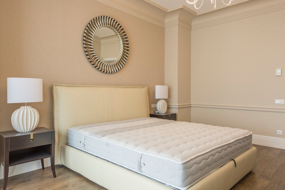 A bedroom interior featuring a large, cream-colored upholstered bed with a high headboard positioned centrally against a pale beige wall. The bed's mattress is plain and slightly textured, with no bedding or pillows visible. On either side of the bed, there are dark wooden bedside tables, each topped with a modern, white ceramic table lamp that has a rounded, ridged base and a simple cylindrical lampshade. Above the bed, a round mirror with a decorative, striped frame in shades of black, white, and beige hangs on the wall, reflecting part of the ceiling and walls. The room is illuminated by soft, natural light, creating a neutral, uncluttered ambiance. The wall has crown molding and a horizontal chair rail, adding subtle architectural detail. The overall aesthetic is minimalistic and elegant, with neutral tones and clean lines, typical of a furnished bedroom awaiting further personal touches. This peaceful setting is typical of a private residence or a well-maintained guest bedroom, aligning with professional waste management services like those offered by Rubbish Clearance Mayfair when arranging for the disposal of unwanted items such as old mattresses or furniture.