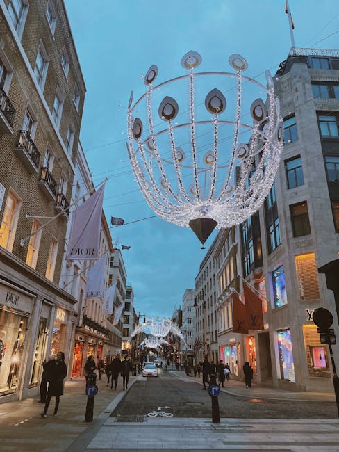 A busy city street during the evening is decorated with large, illuminated chandelier-style festival lights hanging overhead. The chandelier features a spherical frame with multiple circular and teardrop-shaped elements, adorned with numerous small white lights that create a sparkling effect. Surrounding multi-storey buildings on either side display retail shopfronts, including a prominent Dior store with illuminated window displays and signage. Pedestrians, dressed in coats and winter clothing, walk along the pavement, some browsing shop windows and others engaged in conversation. The street is marked with a designated bicycle lane, visible through painted symbol markers, and is lined with bollards to separate pedestrian pathways from the road. The overall scene captures an atmospheric, urban environment with festive lighting and active foot traffic, typical of an alternative approach to city street decoration that complements local rubbish removal and waste management services by maintaining a clean and attractive public space during busy shopping periods.