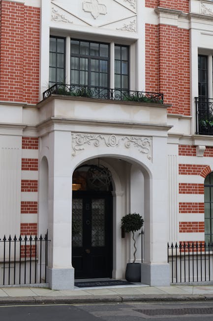 The image shows the entrance to a residential building with a classic architectural style, featuring red brickwork and white decorative stone details. The black door is framed by a large, rounded archway with ornate carvings above it, and it has a vertical window panel with decorative metalwork. To the right of the door, a tall potted topiary with a rounded, dense foliage sits on the pavement. Above the entrance, there is a large window with multiple panes and a small balcony directly below, adorned with black wrought iron railings and green plants in flower boxes. The surrounding façade includes detailed stone embellishments and a symmetrical arrangement of windows and decorative elements. The building appears well-maintained, situated along a quiet street with a small, decorative black metal fence surrounding the pavement area. Rubbish Clearance Mayfair's presence in the context of this image could imply on-site clearance or private disposal in contrast to council-provided waste collection, with the pristine condition of the building suggesting a professional and unobtrusive approach to waste removal at such premises.