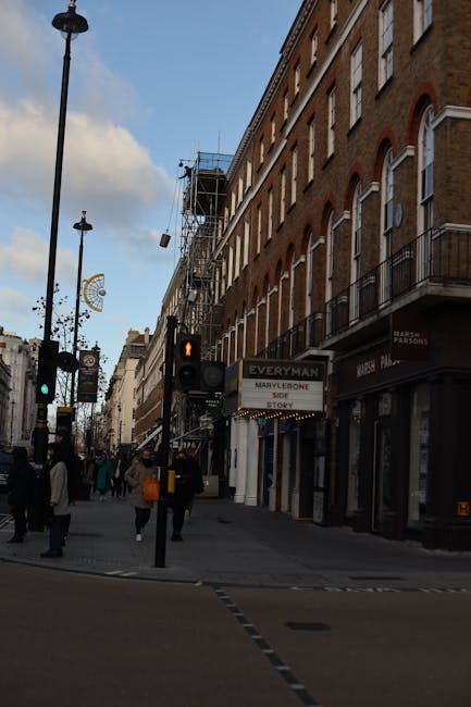 The exterior view of a multi-storey red-brick building with ornate architectural details, including wrought iron balconies and stone embellishments. In front of the building, a black metal fence surrounds a small garden area with pink flowers and green bushes. A traffic light with red signals is positioned on the street corner to the left. Hanging from the building’s façade are two large Union Jack flags attached to poles, extending outward above the entrance, which features a black awning with the name 'Claridge's' visible. Parked along the street are a black taxi and a small blue vehicle. The environment appears to be an urban area, possibly in central London, with a clear daytime atmosphere. This scene exemplifies a typical Mayfair setting, where private property and external decorations are prominent, aligning with independent waste disposal or rubbish clearance services often needed in such locations managed by companies like Rubbish Clearance Mayfair.