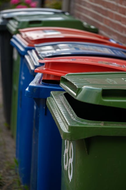 A row of large, wheelie waste bins lined up along a pavement next to a brick wall, with the nearest bin in green having a ridged, textured lid and a white recycling symbol partially visible on its side. Further along, there are additional bins in red, blue, and dark green, each with matching lids, positioned closely together. The bins appear to be made of plastic with smooth, matte finishes, and are designed for outdoor rubbish collection. The scene suggests an outdoor area used for waste disposal, likely part of a private or council-managed collection service, with a natural daylight setting and a blurred background indicating the presence of greenery or other urban elements. These bins are part of an alternative waste handling system, consistent with services like those offered by Rubbish Clearance Mayfair for efficient rubbish management and on-site clearance. The image captures a typical street-side rubbish storage setup, emphasizing the type and organization of waste containers used for household or commercial rubbish collection.