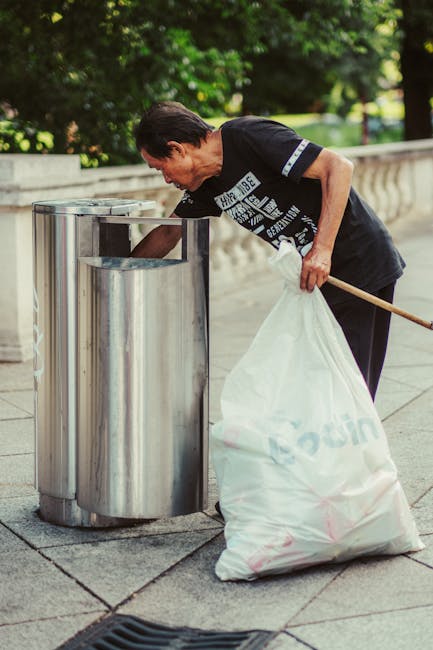 A middle-aged man with dark hair, wearing a black T-shirt with white text and dark trousers, is seen depositing waste into a silver, cylindrical public trash bin on a paved pathway. He holds a white plastic bag filled with rubbish in his left hand while leaning forward to place waste into the bin. The background features lush green trees and a stone balustrade, indicating a park or outdoor public space. The scene is well-lit, with natural daylight highlighting the textures of the man's clothing, the metal surface of the trash bin, and the surrounding environment, suggesting a responsible approach to waste disposal in an outdoor setting. This visual aligns with private waste collection or rubbish removal services provided by Rubbish Clearance Mayfair, emphasizing on-site waste handling in a public area.