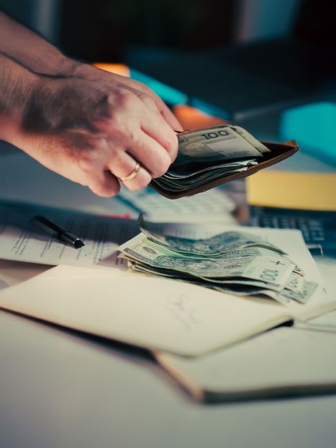 A close-up of a person's hand holding a black wallet, from which multiple banknotes are being pulled out, including a visible €100 euro note on top. The individual is wearing a gold ring on their ring finger. The scene is set on a cluttered desk with various office items, such as pens, a stack of documents, and folders in the background. The lighting is soft but focused, highlighting the hand and currency with a professional, neutral tone. The context suggests financial transactions or cash handling, which may relate to private or alternative waste management payments, akin to consulting Rubbish Clearance Mayfair for independent waste disposal services, though no rubbish is visible in the image itself. The overall composition emphasizes the act of money exchange or management in an office environment, relevant to professional waste management consultations or service quotations.