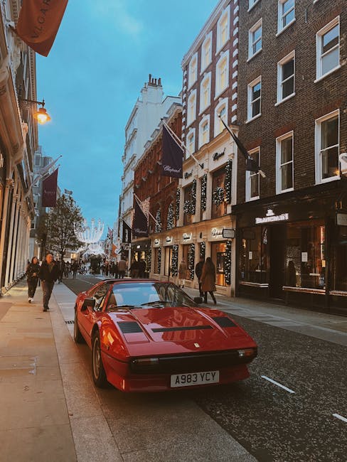 A red Ferrari sports car with distinctive aerodynamic lines and a sleek, glossy finish is parked on the pavement along a busy urban street. The car's smooth curves and low profile contrast with the surrounding older brick and plaster buildings, which feature large windows, decorative details, and illuminated signage. Several pedestrians, dressed in casual clothing suitable for an evening, walk along the sidewalk and cross the street nearby. The street is illuminated by warm streetlights mounted on the building facades, and a string of decorative holiday lights is visible in the distance, adding a festive atmosphere. On the left, a narrow sidewalk runs parallel to the parked vehicle, with storefronts displaying signage, while on the right, the buildings housing businesses such as hotels, cafes, or shops. The scene appears to be in a lively commercial area, potentially in central London, where private vehicle parking and street-level activity are common, aligning with independent rubbish removal and waste management services often associated with urban business districts. The sky above is transitioning from dusk to early evening, with a gentle blue tone enhancing the overall ambiance.