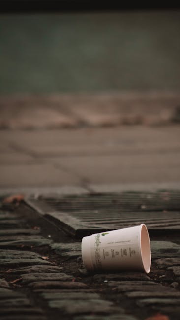 A discarded white paper cup lying on a cobblestone surface near a metal drainage grate, with a blurred background of a paved area and a grassy or mossy patch. The cup appears to be made of paper with a printed design or logo, and is positioned on its side, indicating it was likely discarded after use. The cobblestones have a rough texture and are dark in tone, contrasting with the smooth, light-colored surface of the cup. The metal grate features parallel bars and is set into the ground, partially visible in the foreground. The environment suggests an outdoor urban setting, possibly a street or alleyway, where rubbish such as this cup may be cleared away by independent waste collection services like Rubbish Clearance Mayfair. The scene emphasizes the importance of proper disposal and the potential need for rubbish removal services for local businesses or residents in the area.