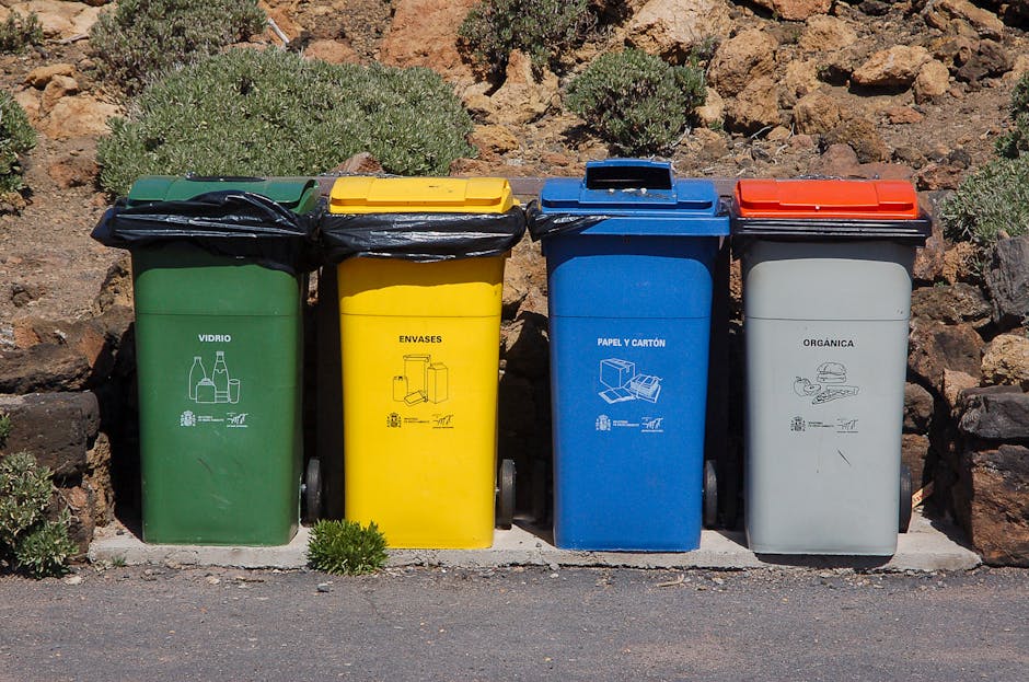 Four wheeled recycling and waste collection bins are aligned side by side on a paved surface, positioned against a background of rocks and sparse shrubbery. The first bin on the left is green with a black lid, indicating it is for glass recyclables, and features a white line drawing of bottles and jars. Adjacent to it, a yellow bin with a matching black lid is designated for packaging materials, with a simple white icon depicting cartons and boxes. Next is a blue bin, also with a black lid, intended for paper and cardboard, marked by a white diagram of folded sheets and boxes. The last bin on the right is gray with a red lid, used for organic waste, illustrated with a white icon of food scraps and garden waste. The setting appears to be an outdoor urban or suburban area, and the presence of the bins suggests a private waste handling or rubbish collection service, such as that provided by Rubbish Clearance Mayfair, instead of local authority collection, supporting alternative waste disposal methods on-site or within a designated waste management zone.
