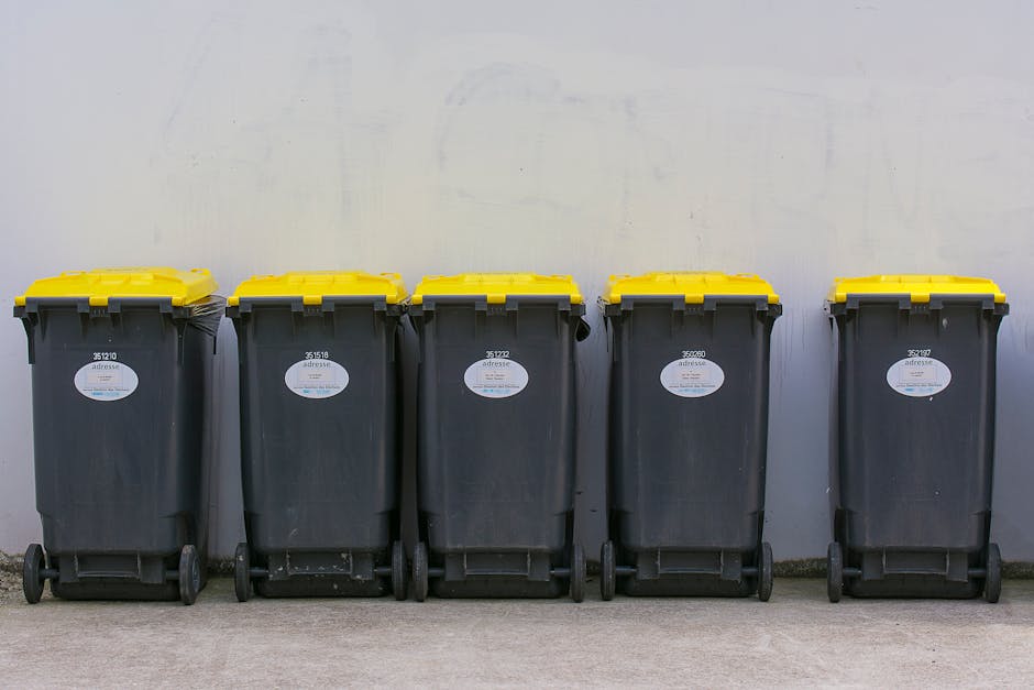 A row of five large wheeled waste bins lined up against a plain, light grey wall outdoors. The bins are made of durable black plastic with textured surfaces, each fitted with a yellow lid securely closed. The bins are evenly spaced, with visible white label stickers on the front displaying identification numbers and barcode details. The ground beneath the bins appears to be concrete, with a smooth finish, and the environment suggests a designated waste storage area, possibly in a residential or commercial setting. The overall scene reflects typical arrangements for private waste collection or rubbish removal services, such as those provided by Rubbish Clearance Mayfair, facilitating efficient on-site waste management and alternative disposal options outside traditional municipal services.