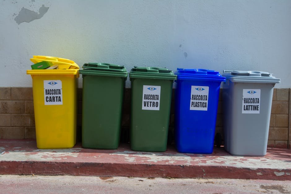 Four wheeled recycling and waste collection bins are aligned side by side on a paved surface, positioned against a background of rocks and sparse shrubbery. The first bin on the left is green with a black lid, indicating it is for glass recyclables, and features a white line drawing of bottles and jars. Adjacent to it, a yellow bin with a matching black lid is designated for packaging materials, with a simple white icon depicting cartons and boxes. Next is a blue bin, also with a black lid, intended for paper and cardboard, marked by a white diagram of folded sheets and boxes. The last bin on the right is gray with a red lid, used for organic waste, illustrated with a white icon of food scraps and garden waste. The setting appears to be an outdoor urban or suburban area, and the presence of the bins suggests a private waste handling or rubbish collection service, such as that provided by Rubbish Clearance Mayfair, instead of local authority collection, supporting alternative waste disposal methods on-site or within a designated waste management zone.