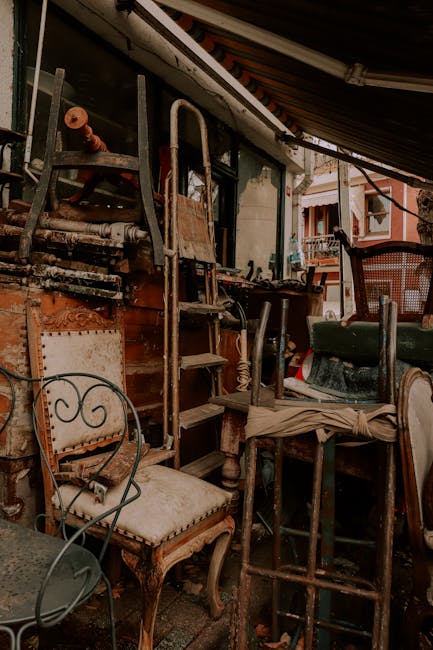 The image depicts an outdoor area cluttered with discarded furniture and miscellaneous items, including a wooden chair with a cushioned seat and a metal chair with a curved backrest, both showing signs of wear. Several wooden and metal objects, such as a small ladder, a stool, and parts of a dismantled chair, are piled together, creating a chaotic scene typical of a clearance or rubbish removal service. The furniture appears aged, with chipped paint and faded upholstery, indicating they are no longer in use. In the background, a wooden structure, possibly part of an outdoor shelter or storage area, surrounds the scene, with a partial view of residential balconies and a shaded awning overhead. The lighting suggests natural daylight, casting shadows and illuminating the varied textures of the mixed materials—wood, metal, and fabric. This scene reflects an environment suited for private waste handling or on-site clearance, with Rubbish Clearance Mayfair capable of managing such large and varied accumulations of rubbish, including old furniture and miscellaneous debris often found in residential or commercial outdoor spaces.