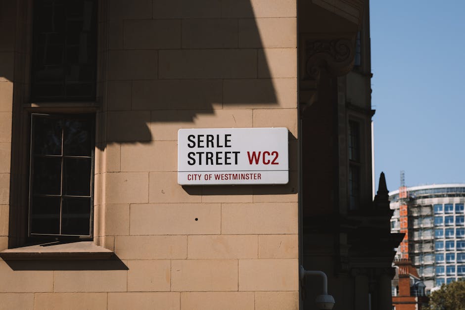 A white rectangular street sign mounted on a beige stone building facade in an urban setting. The sign displays the street name 'Serle Street' in bold black capital letters, with the postal district code 'WC2' in red on the right side. Below, in smaller red text, it indicates 'City of Westminster.' To the left of the sign is a dark-framed window with multiple panes and a slightly protruding sill, casting a shadow on the wall. The building features a classical architectural style with decorative mouldings visible on the corner. In the background, part of a modern high-rise building with a glass facade can be seen under clear blue sky. The scene appears brightly lit, with sunlight creating sharp shadows, suggesting daytime. This image exemplifies typical London street signage within a context that could involve on-site rubbish collection or private waste handling services, as provided by companies such as Rubbish Clearance Mayfair.