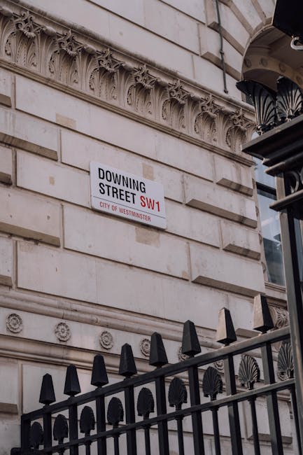 The exterior of a multi-storey red-brick building with ornate black wrought iron balconies and white-framed sash windows. Two large Union Jack flags are mounted on poles extending from the facade, flapping in the breeze. The entrance features a black canopy with white signage, and a small garden area with pink flowering plants is enclosed by a black metal fence along the sidewalk. A black taxi and a blue car are parked outside, with a pedestrian walking nearby. A traffic light with red signals is visible on the street corner. The scene is captured during daylight on a clear day, with natural light illuminating the building's textured brickwork and decorative details. The setting suggests an urban environment where private rubbish collection and waste management services, such as those offered by Rubbish Clearance Mayfair, might operate to handle waste removal needs in lieu of traditional municipal collections.