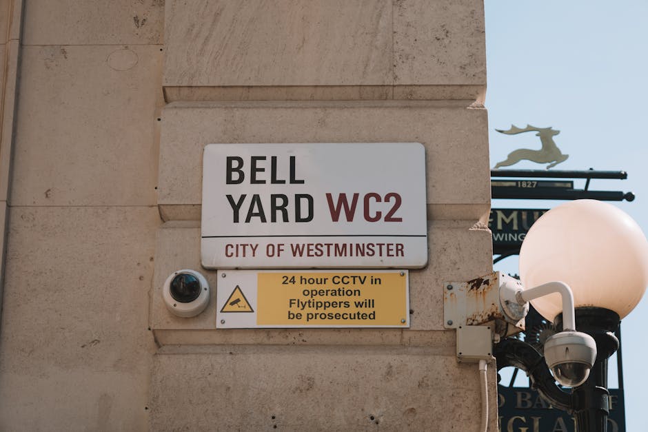 A close-up view of a building corner in Westminster, featuring a rectangular sign with a white background and black and red text indicating 'BELL YARD WC2, CITY OF WESTMINSTER'. Below the main sign, there is a yellow notice warning about 24-hour CCTV monitoring and penalties for flytipping, along with a triangular CCTV warning icon. To the right of the signs, a white security camera with a rounded, dome-shaped lens is mounted on the beige stone wall. Further right, a traditional-style street lamp with a black pole and a large, round, opaque white globe is visible, attached with a decorative arm. In the background, a partially visible blue street sign with a silhouette of a reindeer and some black decorative street furniture can be seen against a clear sky, contributing to the urban environment. The scene depicts elements related to private property and surveillance, aligning with themes of independent waste management and security practices integral to rubbish removal services in Mayfair.