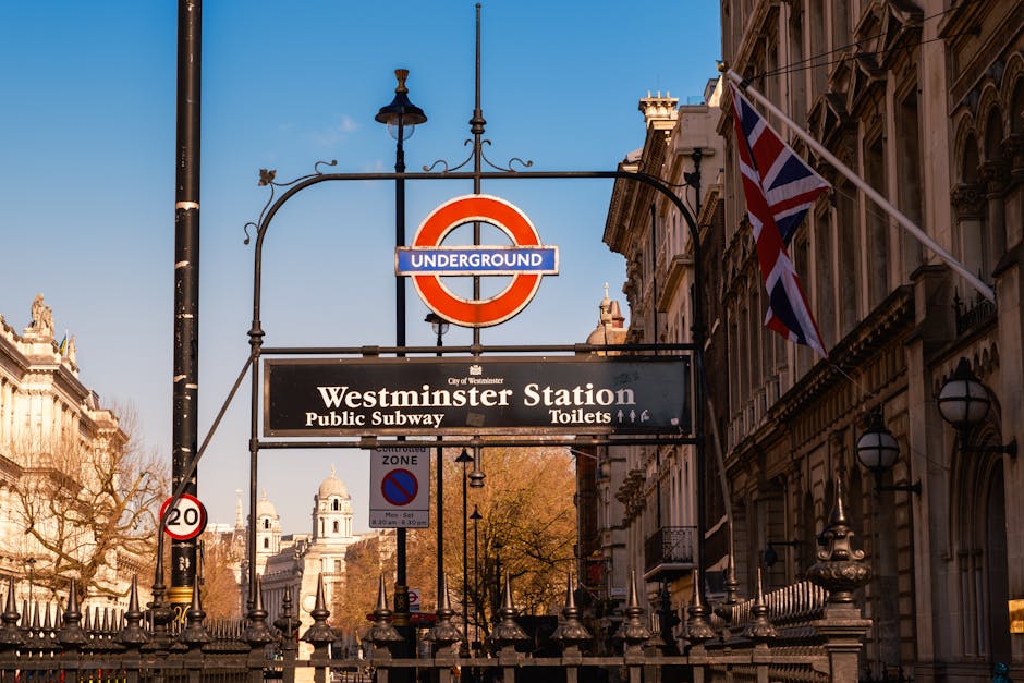 A street scene in front of Westminster Station featuring a prominent underground railway sign with a red circle and blue bar labeled 'Underground', positioned above a black rectangular sign with white text indicating 'Westminster Station' for public subway and toilet facilities. In the background, historic buildings with ornate architectural details and decorative lamps line the street, complemented by a Union Jack flag fluttering on the right side of the frame. The foreground includes black metal fencing with pointed finials, and a small red and white speed limit sign marked '20' attached to a pole on the left. The shot is taken during daylight hours with clear blue sky and warm sunlight illuminating the scene, capturing the urban character typical of central London, relevant to discussions of city infrastructure and waste management near busy transport hubs like Westminster, as referenced on the website rubbishclearancemayfair.com.